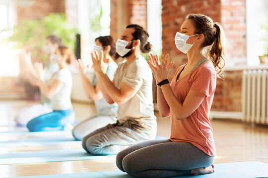 fitness, yoga and healthy lifestyle concept - group of people wearing face protective medical mask for protection from virus disease doing lotus seal gesture and meditating in seated pose at studio