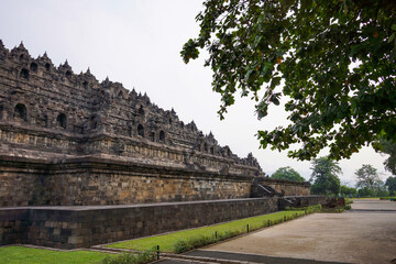Fototapeta na wymiar Exterior wall of ancient Borobudur temple view from the base of temple with trees in foreground and background. No people. Popular tourist and Buddhist pilgrimage destination.