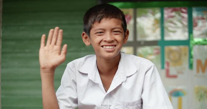 Slow motion scene of a smiling poor Asian elementary school student in white uniform wave his hand for greeting while siting in font of old green wooden wall of his school at the countryside.