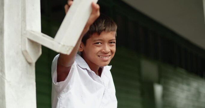 Slow motion scene of a smiling poor Asian elementary school student boy in white uniform siting in font of old green wooden wall of his school at the countryside.