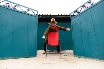 a senior man in a hat with earflaps and a jacket. a pensioner with glasses stands near a red chair that stands near the gate. home owner is home farmer.