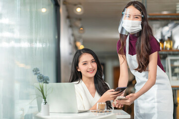 Contactless payment with mobile in restaurant. Waitress standing, wearing protective face mask and apron with asian smiling customer with her smartphone on hand about to pay by qr code payment system