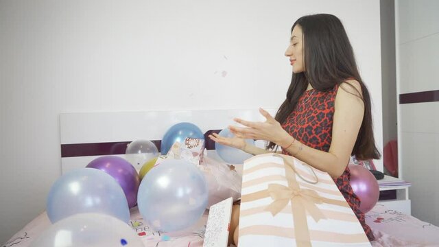 Side view of woman sitting on bed with colorful balloons and opens confetti bomb birthday card