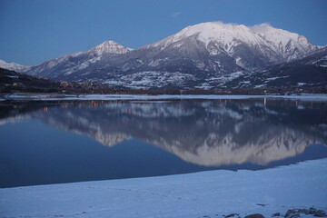 mirror reflection of snow covered mountains, lights from Embrun town at dusk on Serre Ponçon lake, France