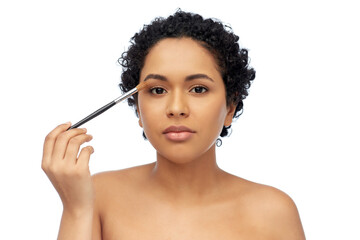 Obraz premium beauty, cosmetics and people concept - beautiful young african american woman with make up brush applying eye shadows over white background
