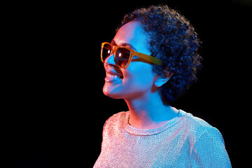 leisure, clubbing and nightlife concept - smiling young african american woman in sunglasses over ultraviolet neon lights on black background