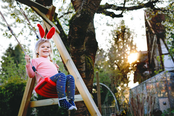 Happy beautiful little toddler girl with red Easter bunny ears having fun on swing in domestic...