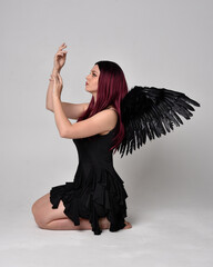 Full length portrait of a red haired  girl wearing black dress and feather angel wings.  Sitting...