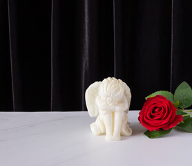 A small sculpture of a baby angel with wings and a red rose on a black background. The concept of funerals, condolences, and mourning.