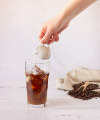 A glass cup with cold coffee and ice, coffee beans on a light background. A hand pours cream and a glass