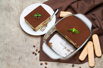 Traditional Italian Tiramisu dessert in glass baking dish, savoiardi cookies and portion on grey concrete background