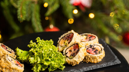 Custom sushi roll in tempura with nori, fresh salmon, tuna, avocado, masago caviar, drizzled with pineapple sauce with salad pouring as decoration on a black plate on a wooden table for Christmas.