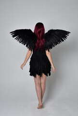 
Full length portrait of a red haired  girl wearing black dress and feather angel wings.  Standing pose against a studio background.