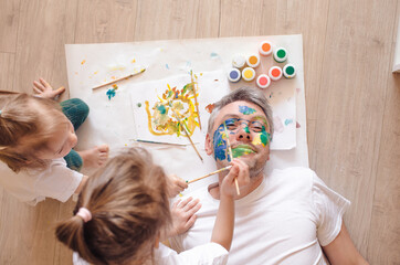 happy dad is lying on the floor, the children paint his face with watercolors. father's day