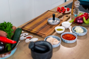 Burger press on wooden cutting board, vegetables and spices with kitchen bowls.