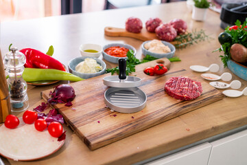 Burger press on wooden cutting board, vegetables and spices with kitchen bowls.