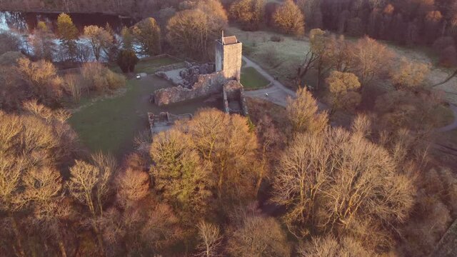Low-level aerial footage over a castle ruin, lit by a low sun, towards a small lake surrounded by trees.