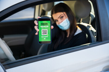 Young woman shows smartphone with international certification of vaccination against Coronavirus Covid-19 infections in car at airport wearing face mask - Health passport concept - Dummy QR code