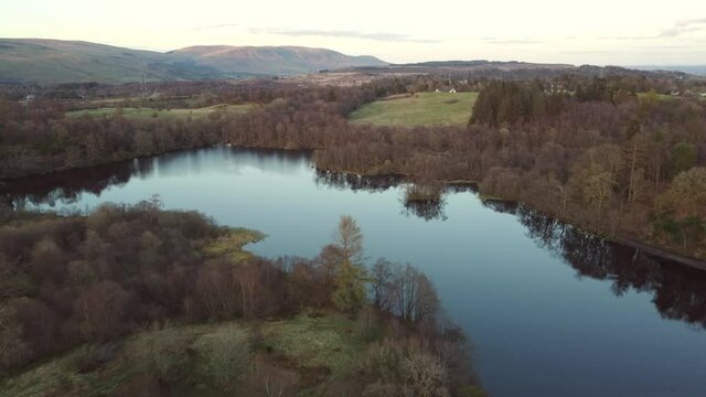 Low-level aerial footage over a small lake surrounded by woodland. With scenic view to hills in the distance.