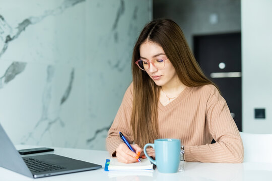 Woman working on laptop at home on kitchen. writing on notepad. Woman trying to find a job. Crysis. Girl styding from home