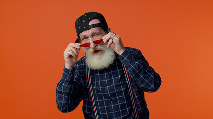 Playful happy elderly bearded man in sunglasses and cap blinking eye, looking at camera with toothy smile, winking and flirting, expressing optimism. Senior mature old grandfather on orange background
