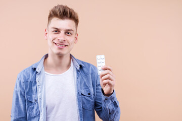 Young man in denim shirt is holding medicine anesthetic, antipyretic, sedative pills over isolated beige background celebrating achievement with happy smile and winner expression. Health care concept.