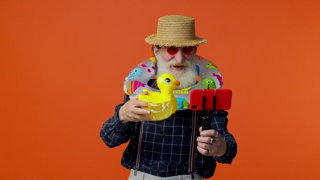 Mature Grandfather Traveler Blogger With Swimming Ring And Inflatable Duck Toy, Taking Selfie On Mobile Phone, Make Video Call Online. Studio Shot. Senior Man On Summer Holiday Vacation, Trip. Tourism