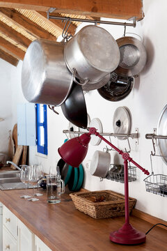 bancada de cozinha r&uacute;stica, com candeeiro de p&eacute; em metal tachos e panelas pendurados em ferro e aluminio, cesta de verga, copos de jarro de vidro com &aacute;gua. cozinha de campo