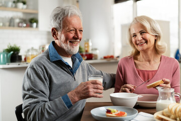 Senior couple eating breakfast in the kitchen. Husband and wife talking and laughing while eating a sandwich.
