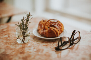 Croissant on a pink marble table with reading glasses and a small branch of rosemary on a small vase