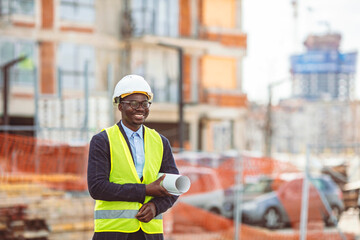 Portrait of African American man architect wearing a vest and helmet, he stands and holding a blueprints with the sky and urban in background. Engineer and architect concept.