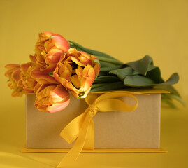 Orange-red fresh tulips in a bouquet and a gift box on a yellow background. Greetings, celebration, romance concept. Copy space. Bright colorful postcard for congratulations on all holidays and events