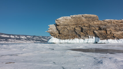 Fototapeta na wymiar A picturesque rocky island rises above an icy lake against a blue sky. A rock with bizarre outlines, layered structure, covered at the base with ice splashes, icicles. Winter day. Baikal