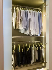 Opened door wardrobe displays many shirts and pants with LED vertical lighting inside.