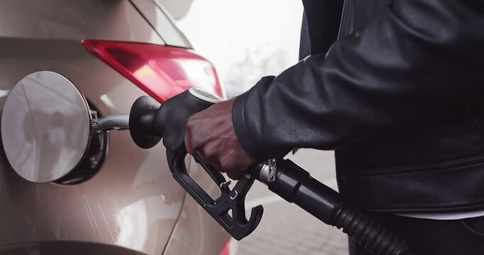 Cropped close up image of unrecognizable African businessman in casual wear, rholding in hand filling gun and refueling his luxury car with gasoline at the petrol station