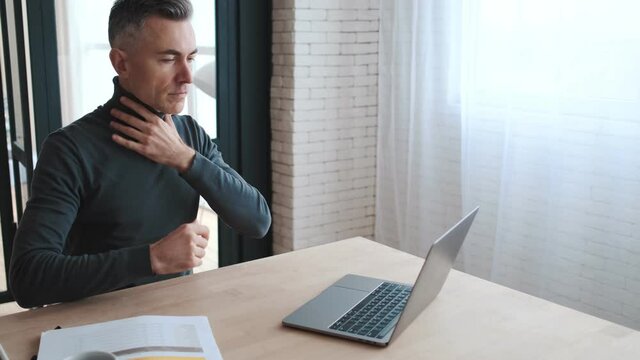 The side view of a mature man in a green turtleneck sitting at a table in front of a laptop with glasses, fixing his hair and revising papers in a room