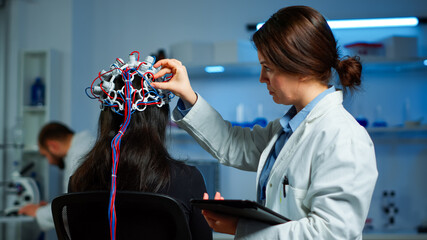 Woman patient wearing performant eeg headset scanning brain electrical activity in neurological research laboratory while medical researcher adjusting it, examining nervous system typing on tablet.