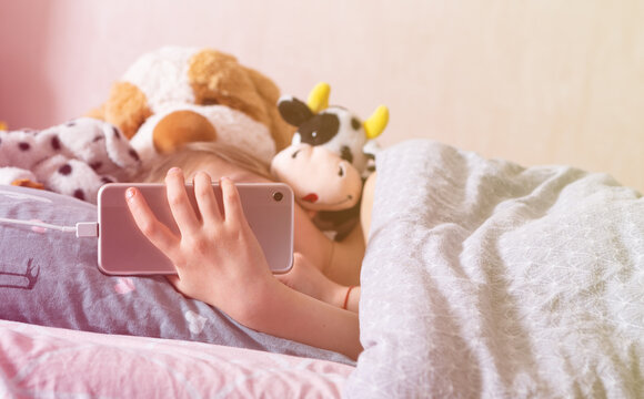in the evening, the child lies in bed and holds a cell smartphone in his hand, next to her are favorite soft toys