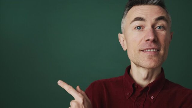 A close-up view of a pleased elder man is pointing to the copy-space standing isolated over green background in the studio