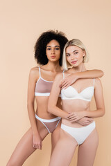 african american woman hugging blonde friend while posing isolated on beige