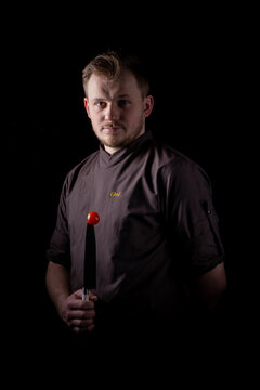 A Chef In A Uniform Jacket Shows Off A Professional Chef's Knife And A Tomato