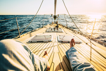 pov point of view of man legs laying and relaxing on the wooden sail boat deck alone with sunset in background - concept of travel people and freedom in summer holiday vacation - obrazy, fototapety, plakaty