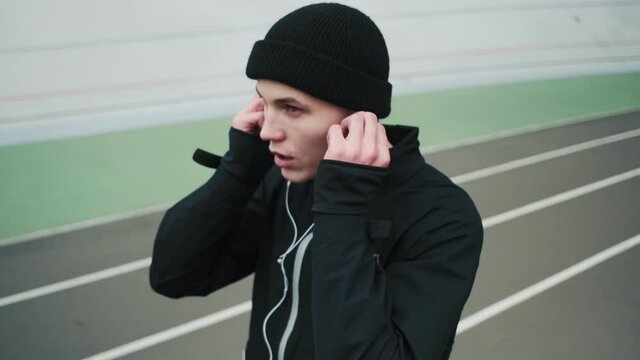 A strong young sportsman guy wearing a black sport-suit is putting on his earphones while training at the stadium