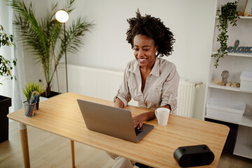 Beautiful black woman is sitting for wooden desk, working and typing on laptop at home office.