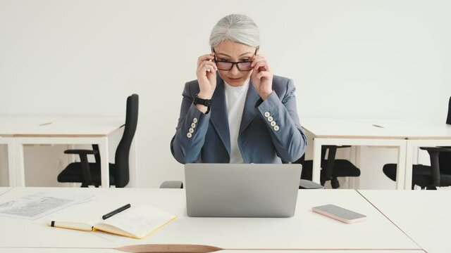 A beautiful elder businesswoman is putting on her eyeglasses while working in the office