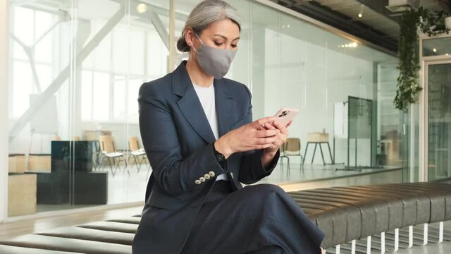 A beautiful elder businesswoman wearing facial protected mask is using her smartphone while sitting in a conference room