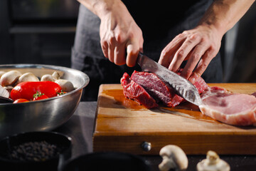 The male chef cuts raw meat with a knife on a wooden board. The restaurant kitchen cooking.