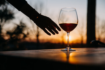 Female hand taking a glass of red wine from a wooden table outdoors in sunset light.