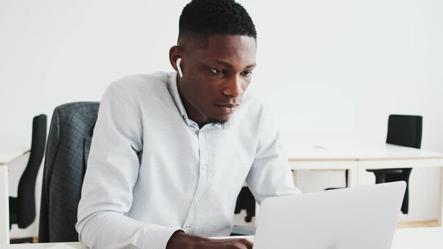 A focused black guy with earbuds is using his laptop while working in the office