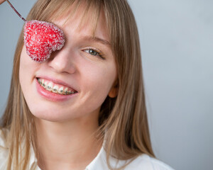 Cute woman with braces on her teeth holds a candy in the form of a heart on white background